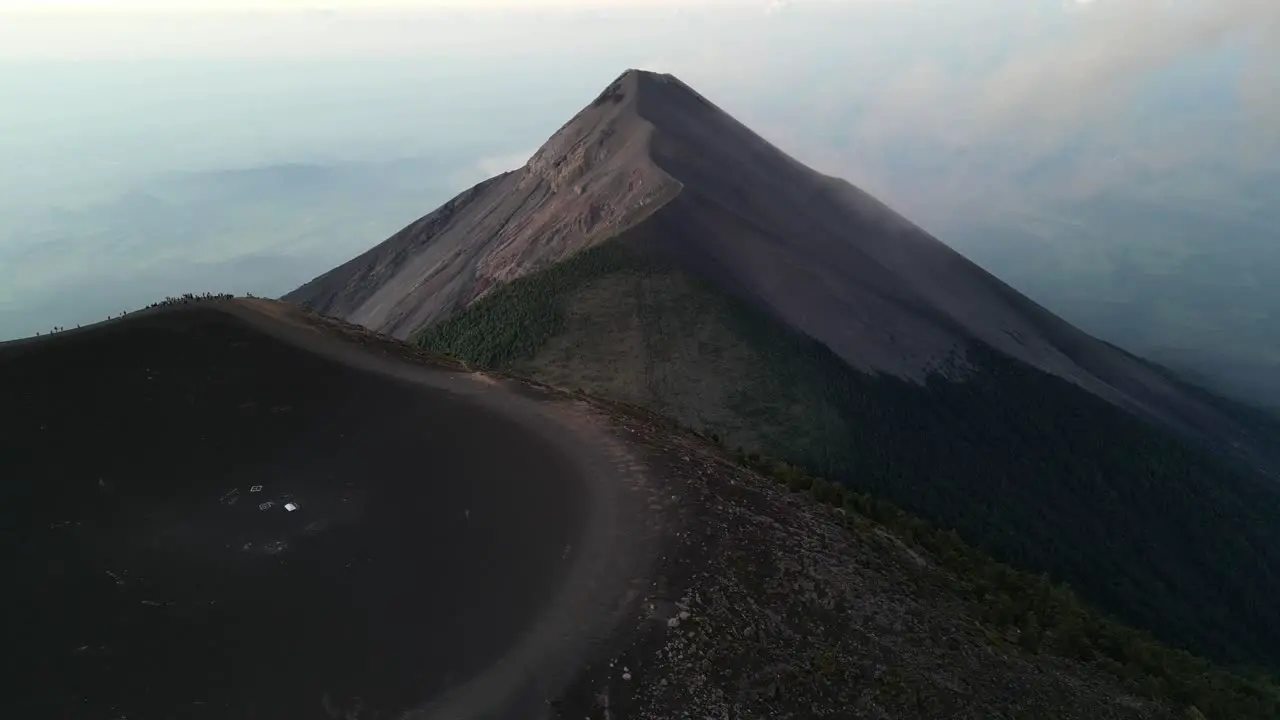 Drone view in Guatemala flying over a volcano crater covered in ashes at sunrise surrounded by green mountains and clouds over towns