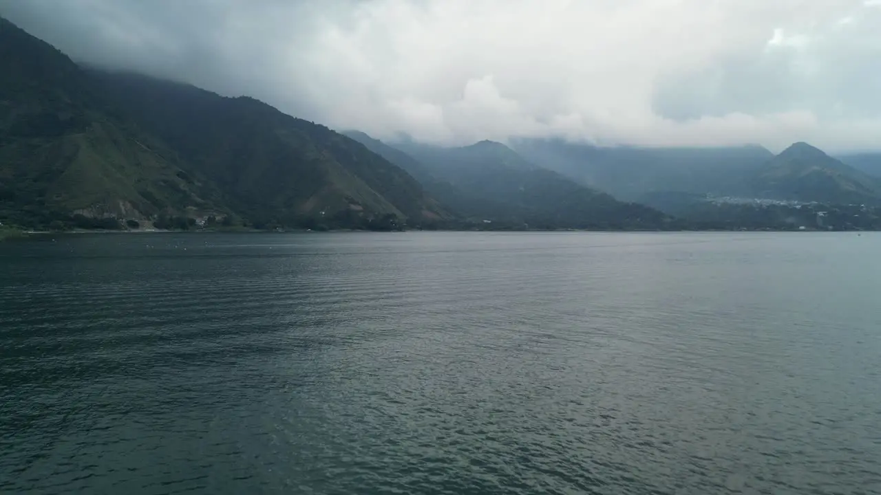 Drone view in Guatemala flying over a blue lake surrounded by green mountains and volcanos on a cloudy day in Atitlan