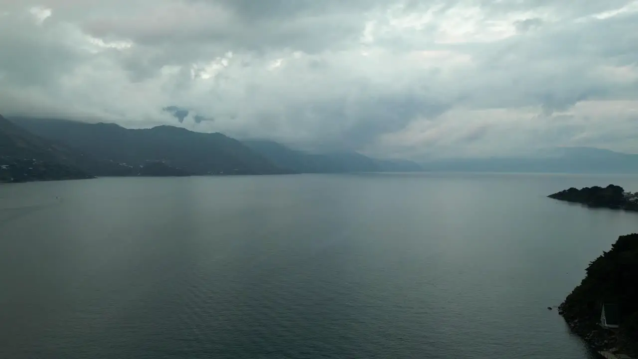 Drone view in Guatemala panning a blue lake surrounded by green mountains and volcanos on a cloudy day in Atitlan