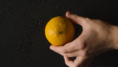 Overhead Studio Shot Of Hand Picking Up Orange With Water Droplets Revolving Against Black Background