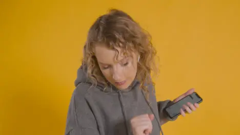 Studio Shot Of Young Woman Listening To Music On Mobile Phone And Dancing Against Yellow Background 1