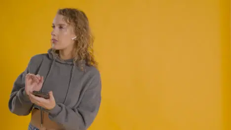 Studio Shot Of Young Woman Listening To Music On Mobile Phone And Dancing Against Yellow Background 2