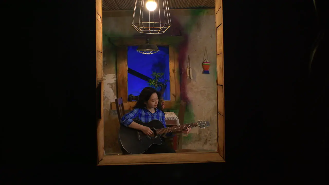 young woman plays the guitar at night as visible through a window