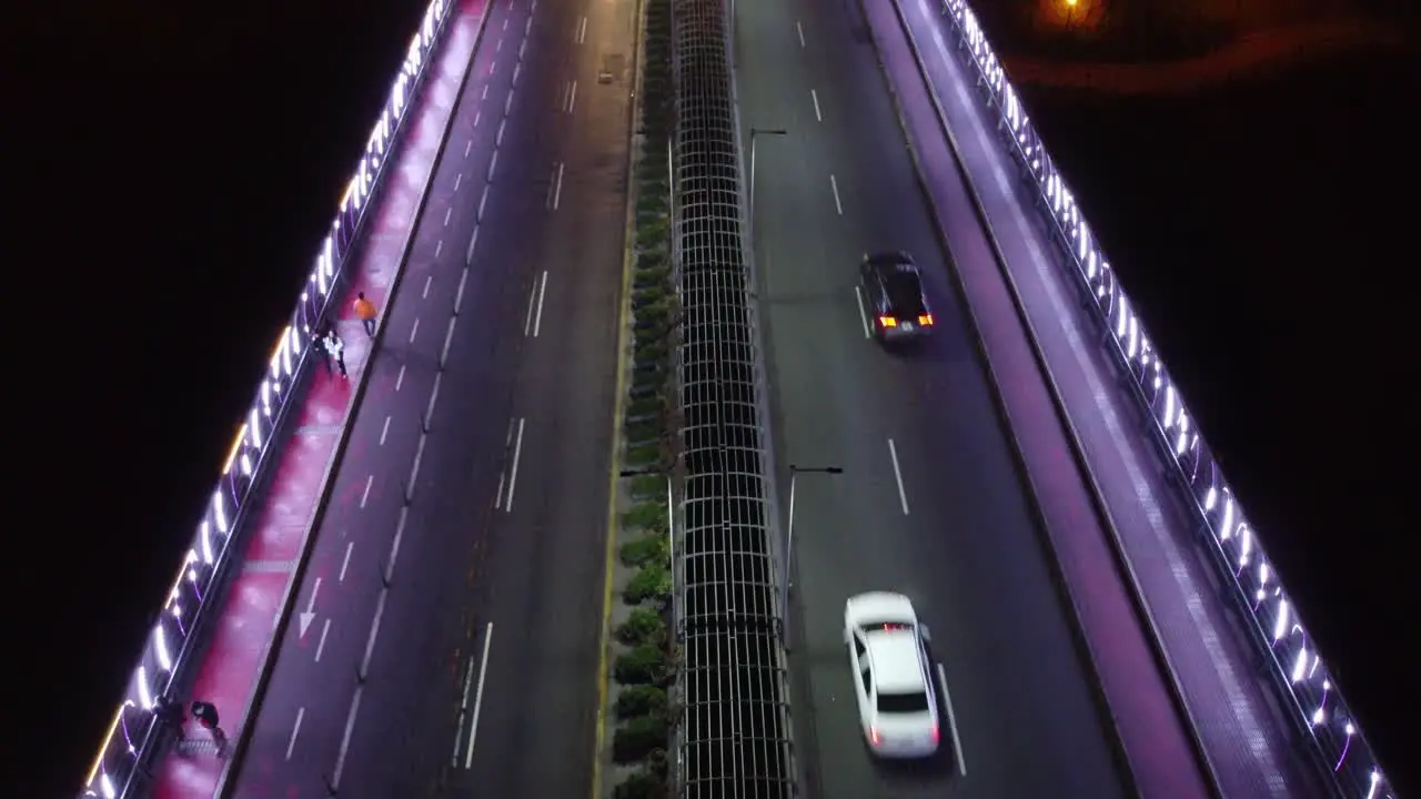 Drone video of a bridge at night lit with purple lights People walk by on sidewalk as cars drive by in the street lanes