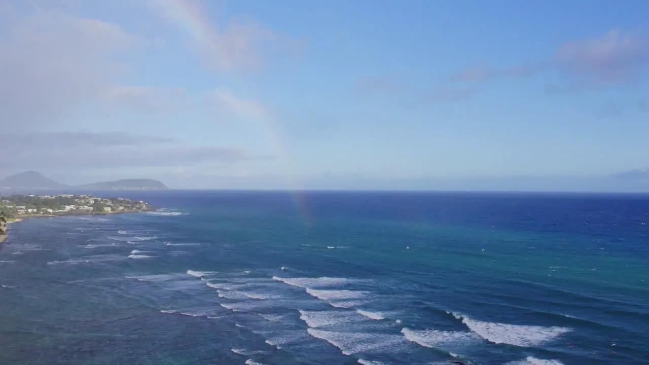 Drone footage catches a rainbow in the ocean mist along the coast of Oahu in the Hawaiian islands with volcanic mountains on the horizon and white capped waves rolling onto the shore