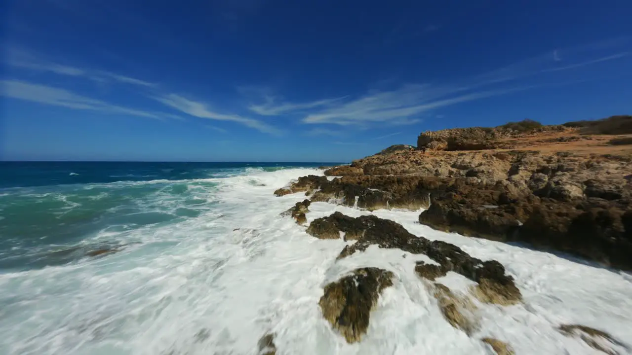 FPV Racing Drone over crystal clear blue splashing water flying towards rocky coast line shore line