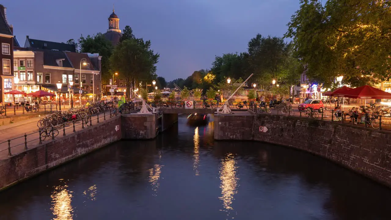 A wide timelapse of a busy intersection with pedestrians cyclists traffic and boats on a canal in nighttime Amsterdam