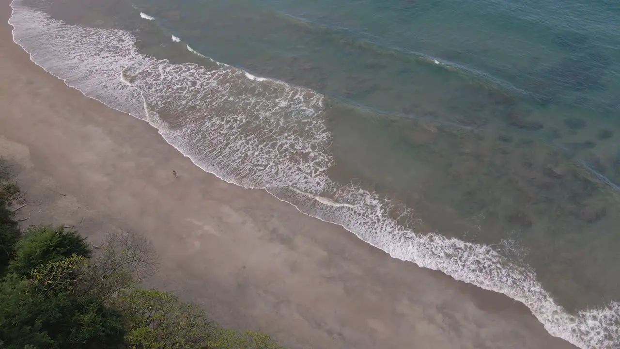 Descending Aerial Tracking Shot Of A Young Woman Walking Leisurely Across A Beautiful Sandy Beach