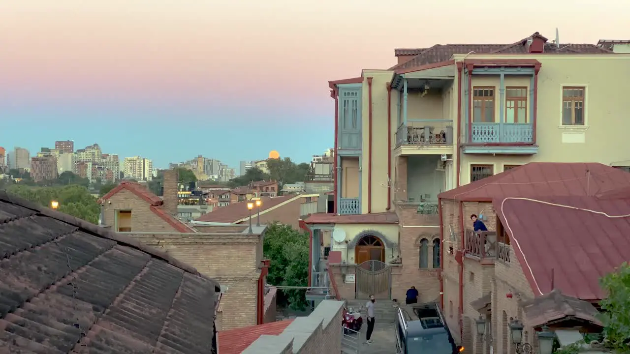 Moon rise over Tbilisi Georgia in the high mountain of Caucasus in Europe city center café historical building old house with red tile roof pink blue sky and cobblestone walkway couple waling together