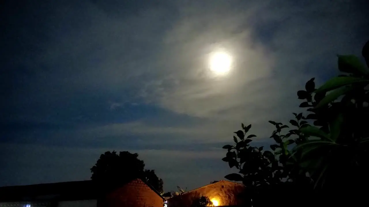 Moon motion lapse from left to right with visible house roofs and plants on slightly cloudy night