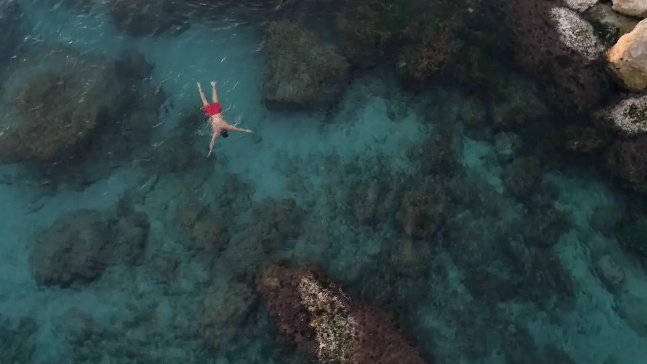 Young Caucasian man swims in gorgeous clear turquoise water by large rocks circle aerial shot