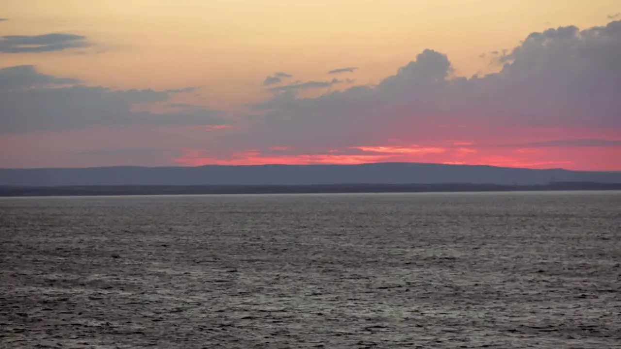 View from sea of sun setting behind clouds and hills near Sydney Nova Scotia with beacon flashing on shore