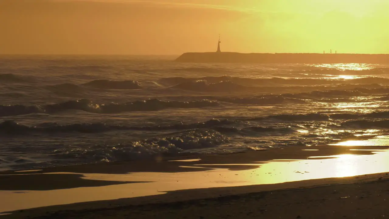 Rough seas at sunrise waves rolling onto sandy beach with harbor wall in background