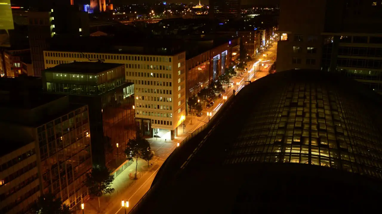 A high-angle timelapse unveils a night cityscape foregrounded by a large lit glass-roofed building set against a backdrop of glowing street-lit streets and illuminated buildings