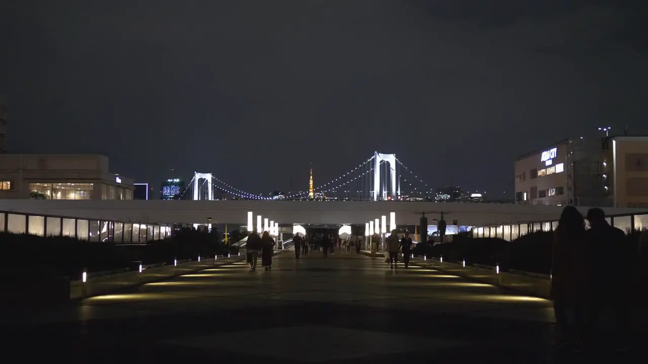 Night scene of Odaiba walkway with people walking by and the Rainbow Bridge in Tokyo Bay in the background Tokyo Japan