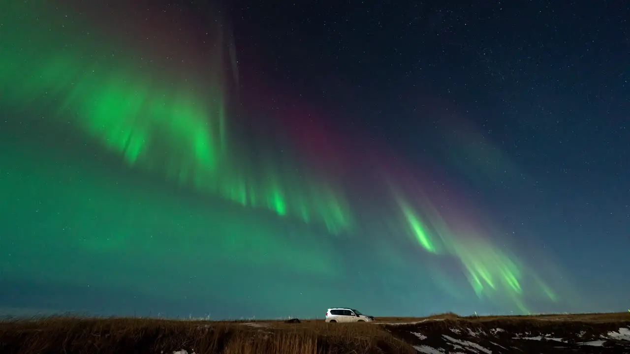 Time lapse shot of flickering green and purple aurora borealis at night sky of Iceland with parking car on road wide shot