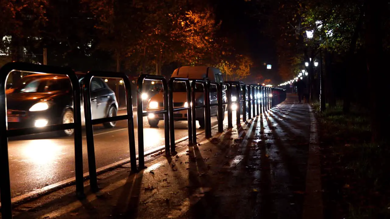 Cars moving slowly on city street at night lights flare shining through railing irons bordering sidewalk