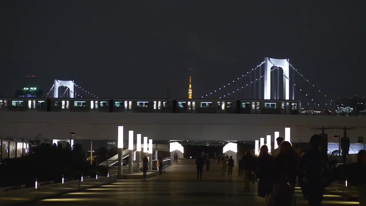 Night scene of Odaiba walkway with people walking by and a Monorail car passing with the Rainbow Bridge in the background in Tokyo Bay Tokyo Japan