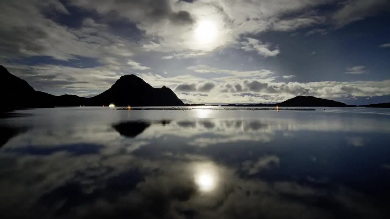 Night scene with the full moon shining behind the floating clouds with their reflections on the calm fjord