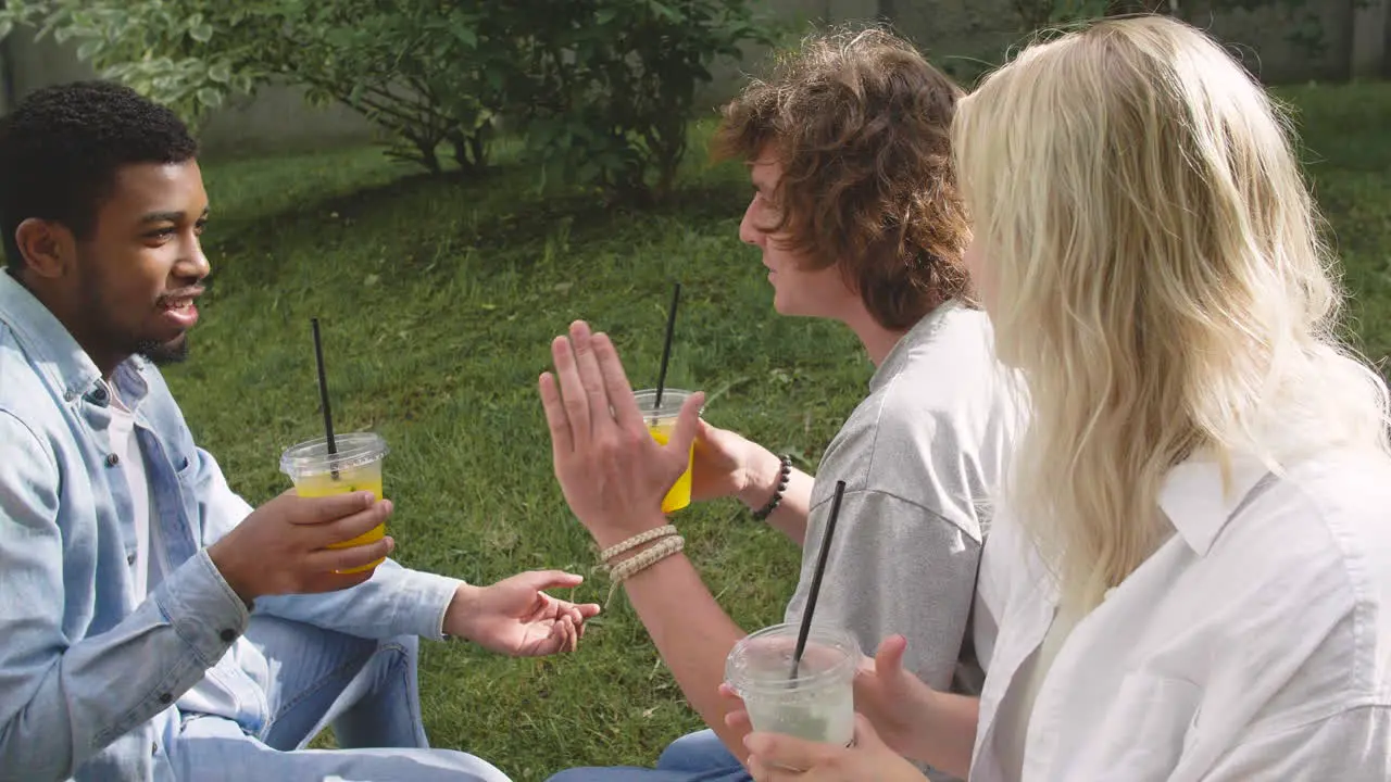 Happy Multiethnic Friends Spending Time Together Sitting In A Park Drinking Refreshing Juice And Chatting