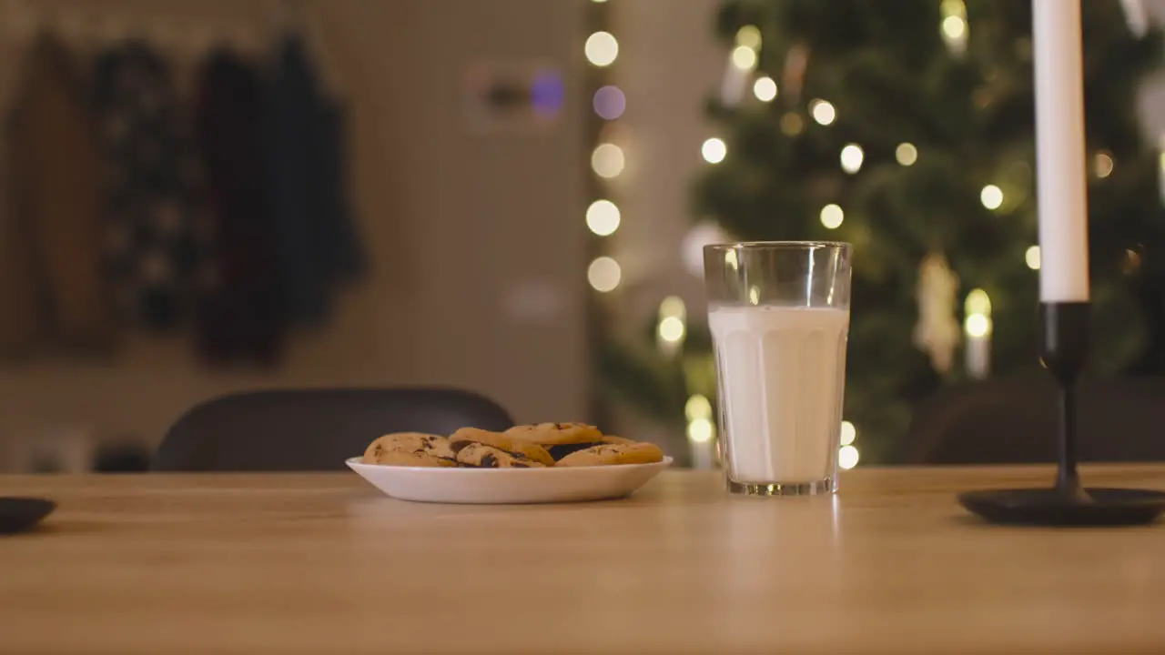 Camera Focuses On An Glass Of Milk And A Plate Full Of Cookies On An Empty Table With Two Candles In A Room Decorated With A Christmas Tree