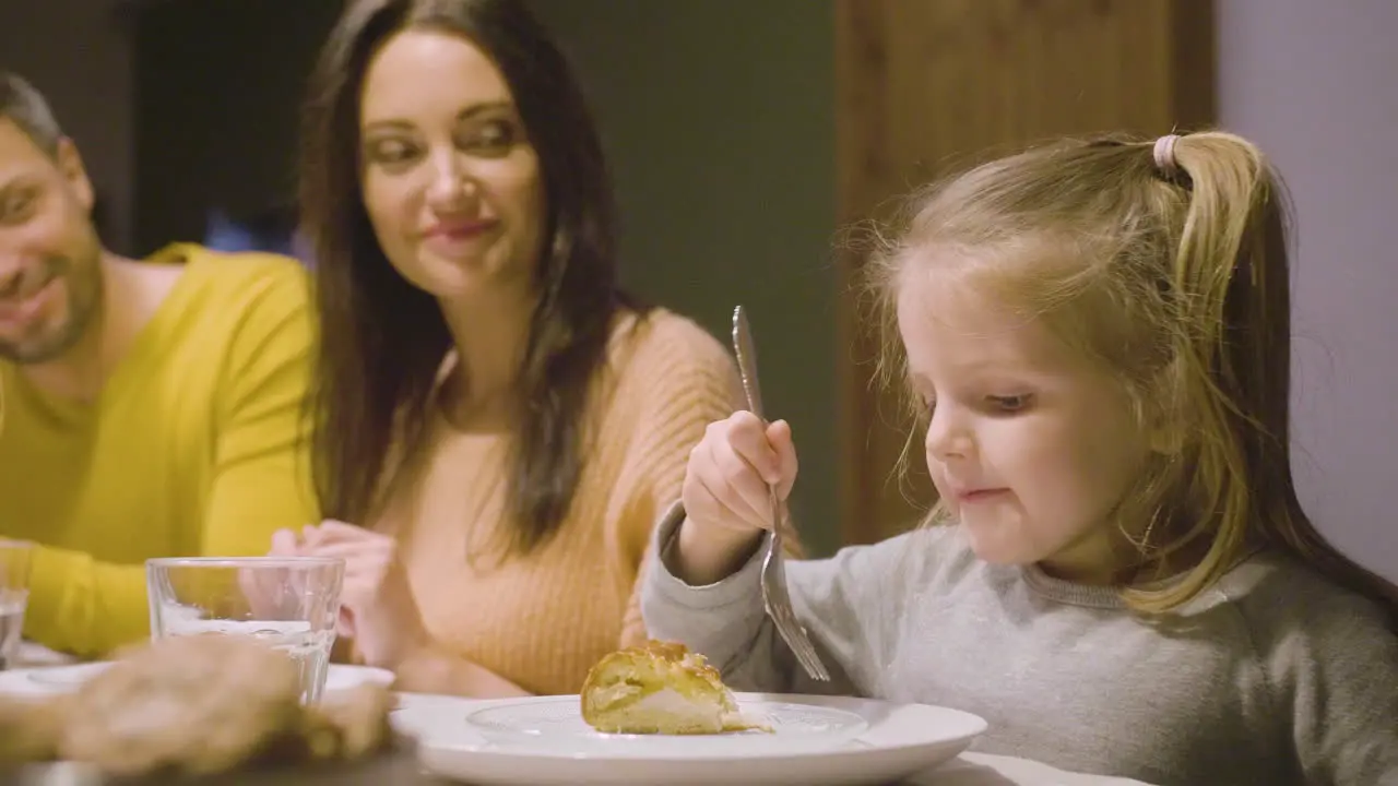 Side View Of Blonde Little Girl Eating Apple Pie Sitting At The Table While Her Parents Watching Her At Dinner