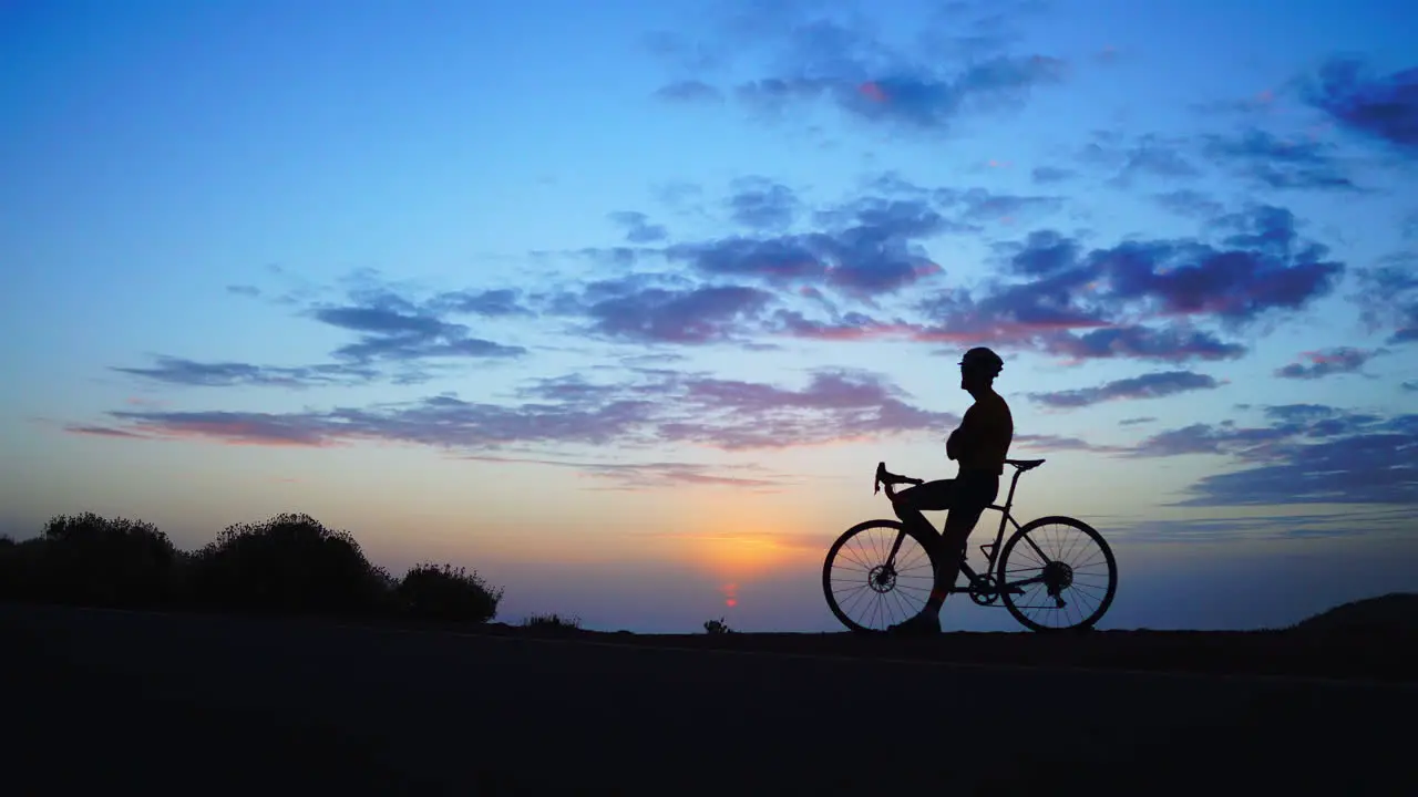 The man stands alongside a bicycle framed against the sunset in the background The moment is captured in a time-lapse sequence using a wide-angle view