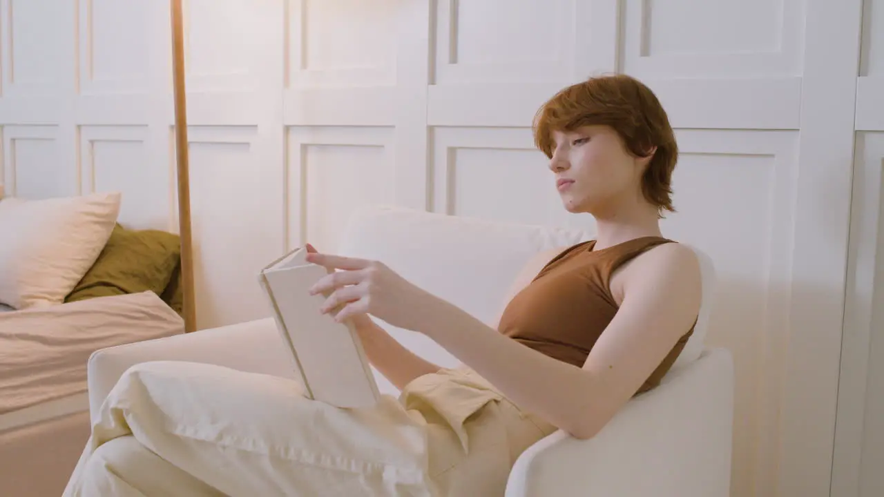 Side View Of Girl Sitting On A Chair Reading A Book In The Bedroom