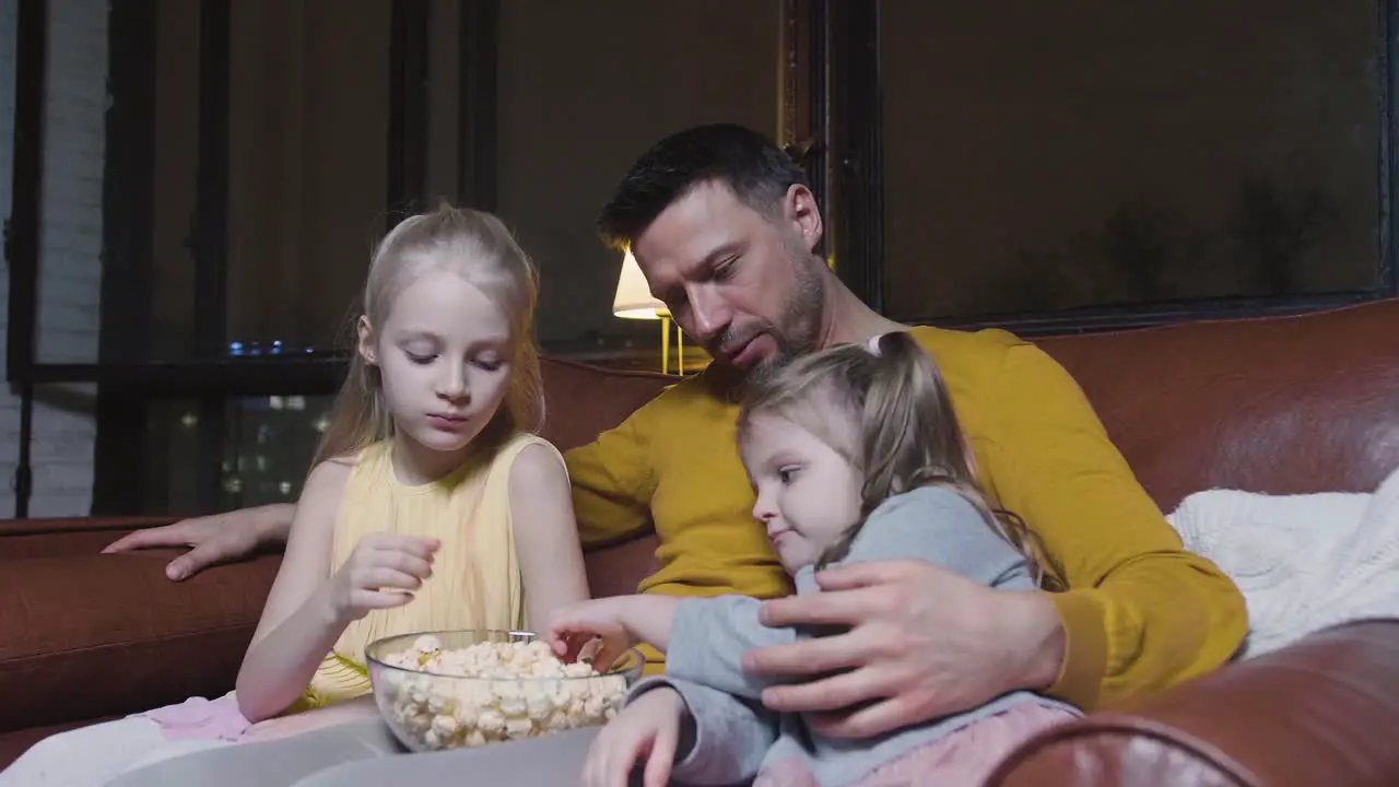 Dad And His Two Little Daughters Eating Popcorn And Watching A Movie While Sitting On Sofa At Night At Home