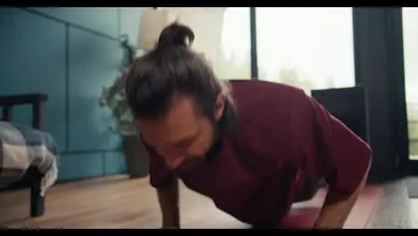 A happy brunette guy in a red t-shirt has done the push-up exercise at home gets up on his feet and wipes his forehead Fitness at home