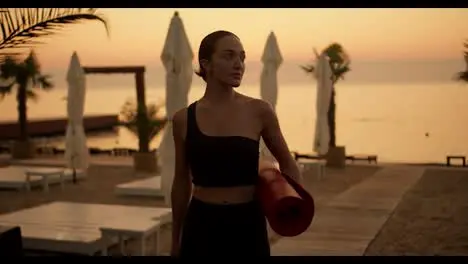 A middle-aged girl in a black tracksuit with a red mat in her hands leaving the beach after an evening workout and yoga class Golden hour Golden sunset on the beach