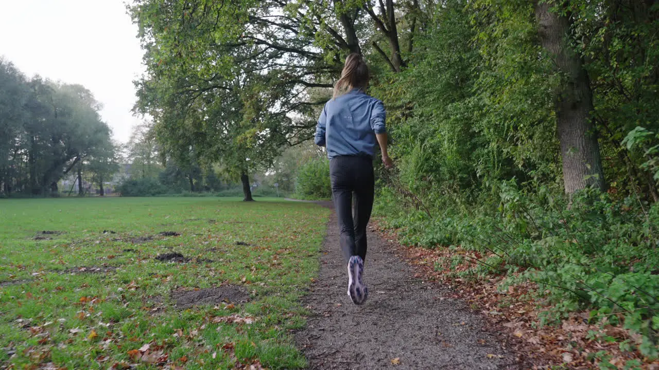 Dynamic slow motion shot camera follows active woman running through park on path forest on right grassfield on left