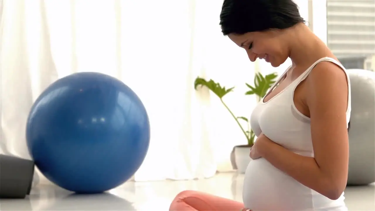 Smiling pregnant woman sitting on mat touching her belly