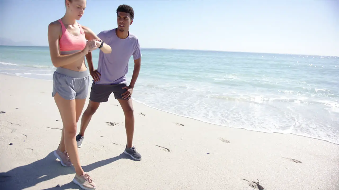 Young Caucasian woman checks her smartwatch beside a young biracial man on a sunny beach with copy s