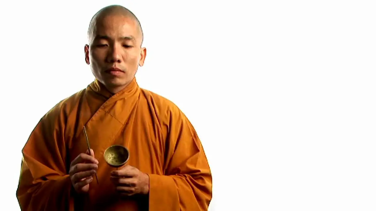 A Buddhist monk wearing an orange robe taps a bell with a small stick