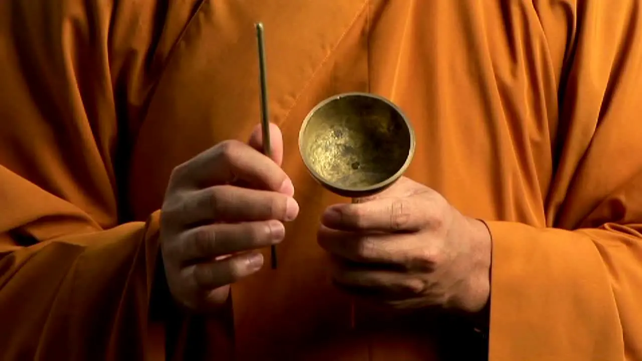 A Buddhist monk wearing an orange robe taps a bell with a small stick 1