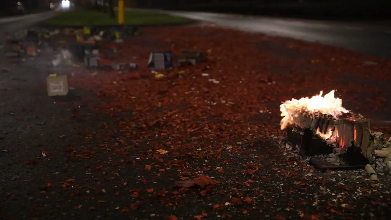 Flame on fireworks burn out in the middle of the street as cars drive by and debris fills the roadway