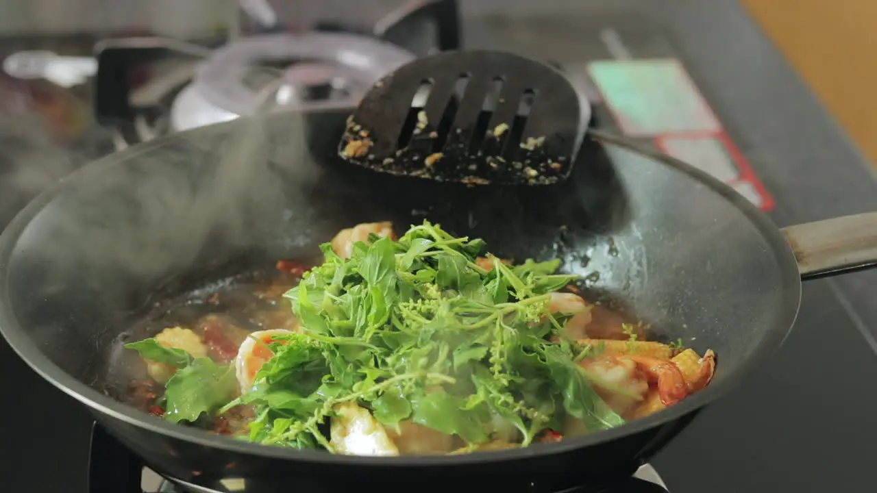 Girl is adding fresh bunch of green basil leaves to frying shrimps