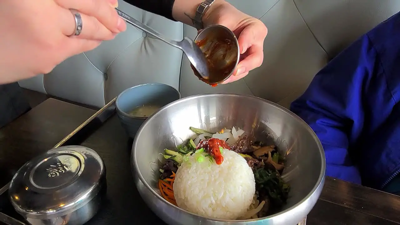 2-3Korean food served and mixed by waitress to lady dining in at modern futuristic resturaunt with contemporary articulated metalic utensils bowls and seated at a lit white pleather bench wooden table