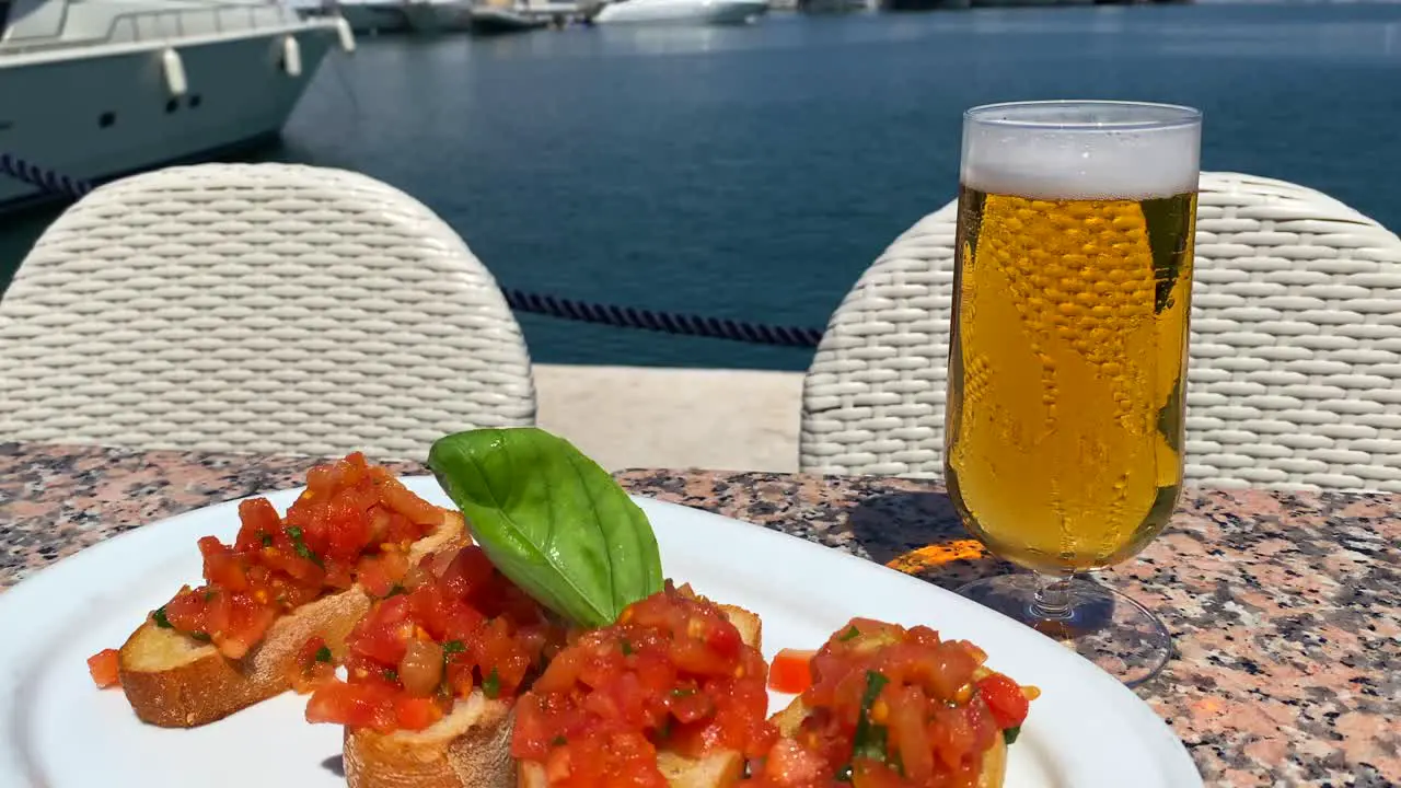 Classic traditional Italian bruschetta bread with tomatoes and olive oil cold beer glass and view on luxury yachts in Puerto Banus port during summer in Marbella Spain tasty food 4K shot