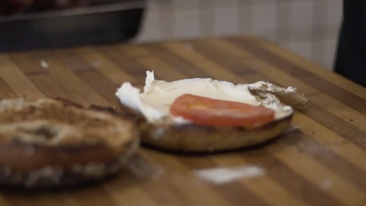 Close up shot spreading cream cheese and adding tomatoes onto bagel