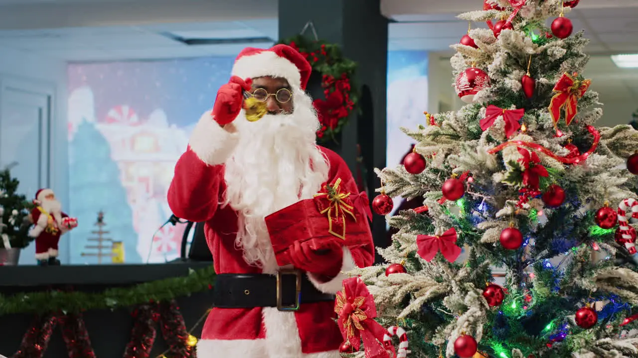 African american employee posing as Santa Claus ringing bells next to beautifully decorated Christmas tree trying to attract customers to fashion store during promotional holiday season