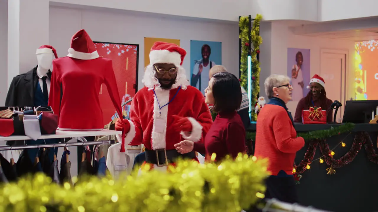 Employee wearing Santa Claus costume around shopping mall clothing shop to spread holiday joy Worker in festive themed suit showing happy client red garment piece during winter holiday season