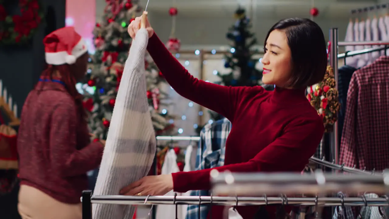 Asian client browsing through rack of clothes in Christmas themed shop checking for fitting blouse size Woman in xmas ornate fashion boutique during festive holiday season