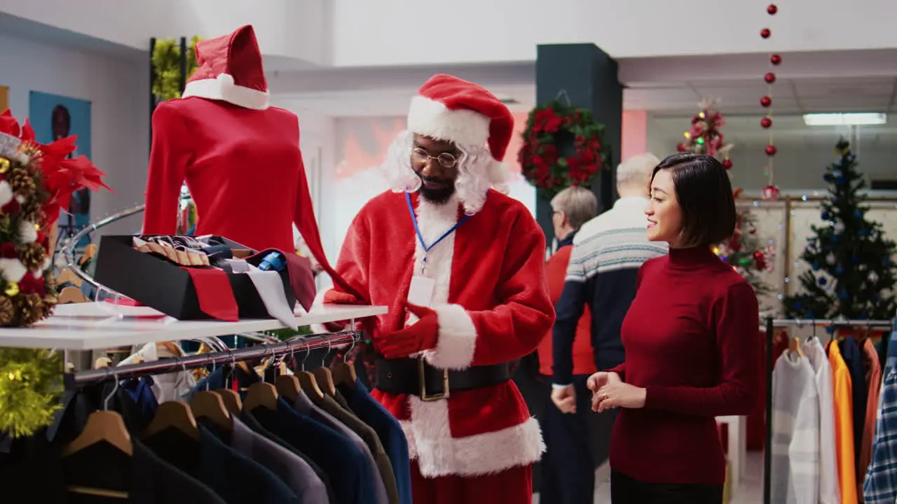 Retail assistant wearing Santa Claus costume around shopping mall clothing store to spread holiday joy Employee in xmas themed suit showing happy customer red garment piece