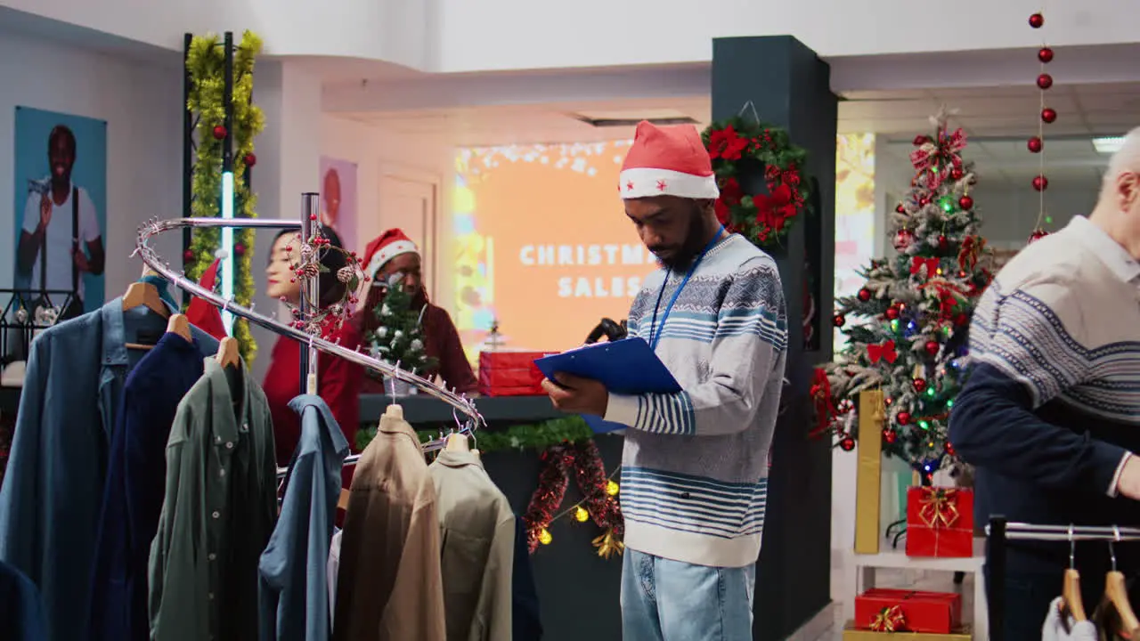 Manager wearing Santa hat and holding clipboard browsing through clothes racks at Christmas themed clothing store sorting out price tags during holiday season promotional sales