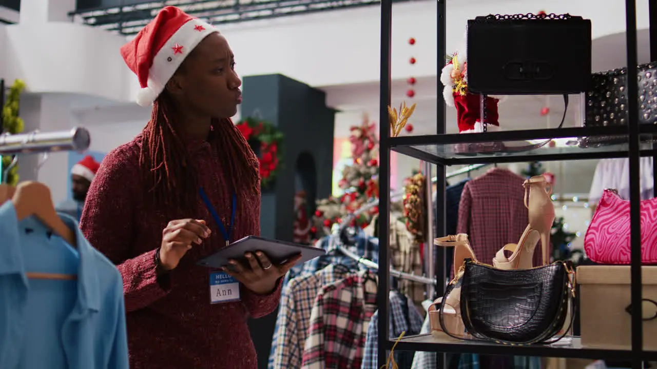 African american supervisor wearing Santa hat browsing through clothes racks at Christmas themed fashion boutique using tablet to input promotional prices on online store during holiday season sales