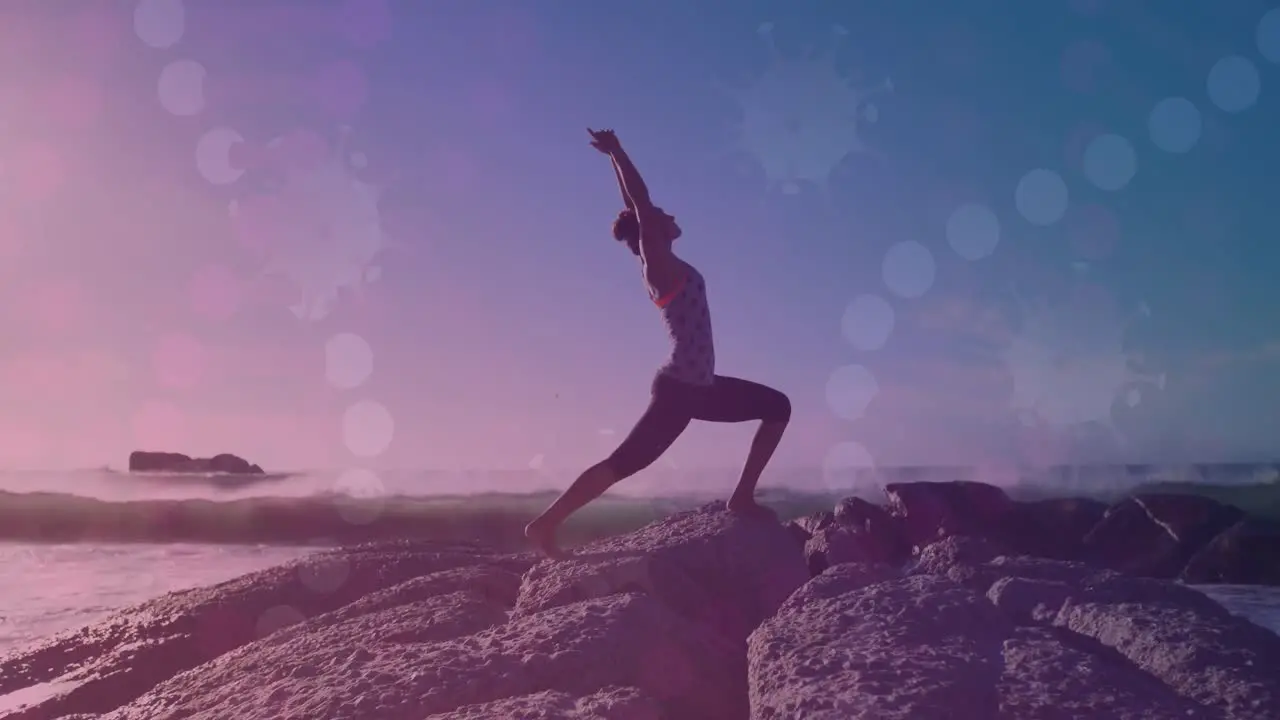 Animation of glowing lights over biracial woman practicing yoga by the sea