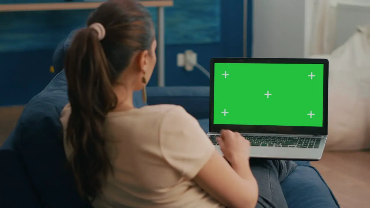 Freelancer woman sitting on sofa in home office typing on laptop computer with mock up green screen chroma key