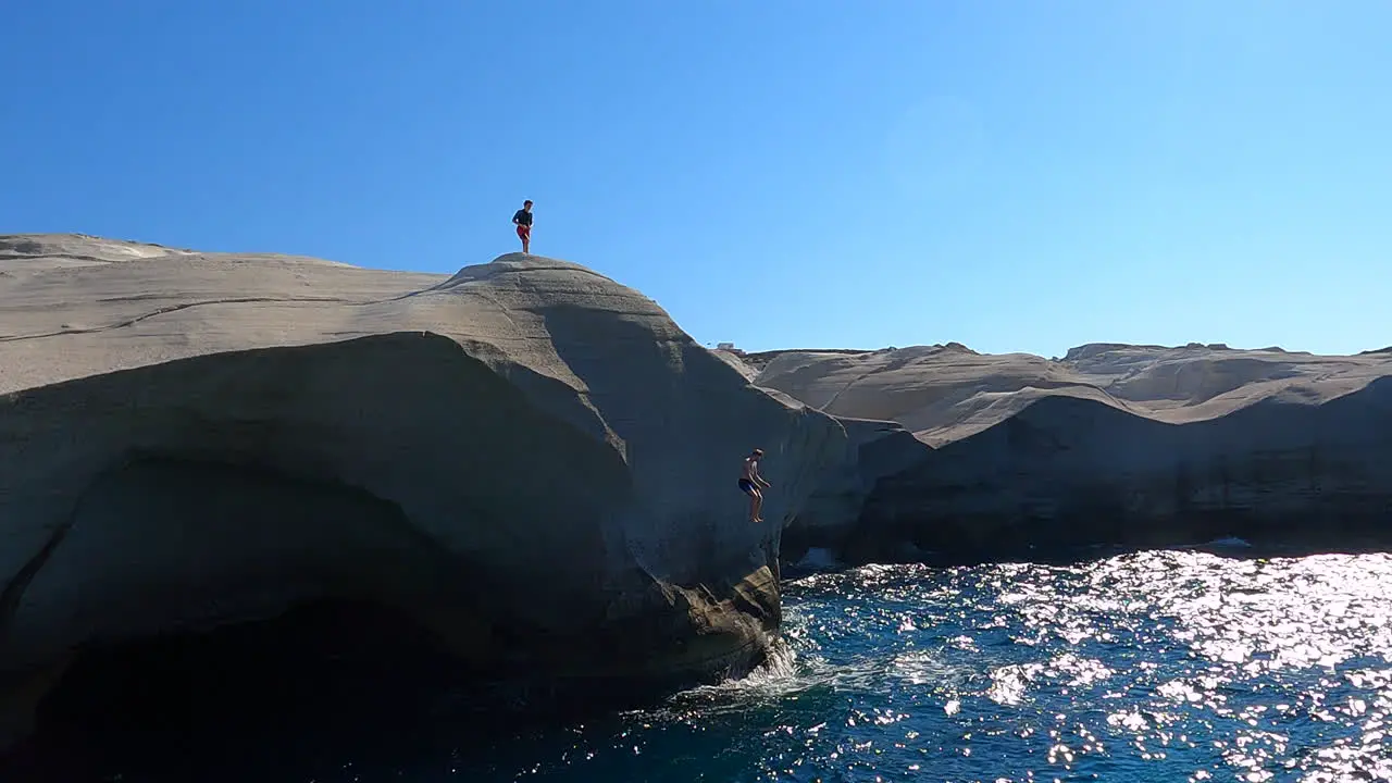 Two people stand on the highest point of a cliff one jumps into the water of the Mediterranean sea